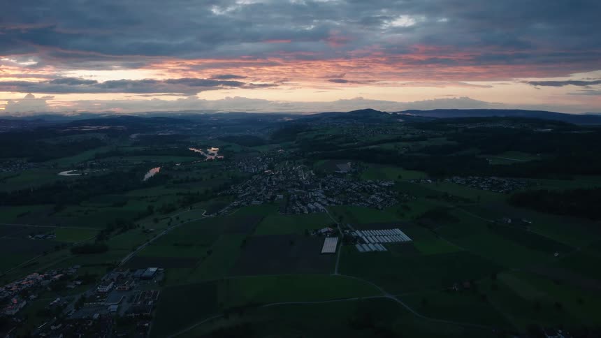 An expansive aerial shot captures a peaceful Swiss village and surrounding green fields during a beautiful, moody sunset
