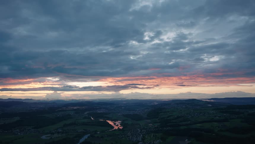 A dramatic cloudy sunset paints the sky pink and orange over a rolling green valley with a winding river