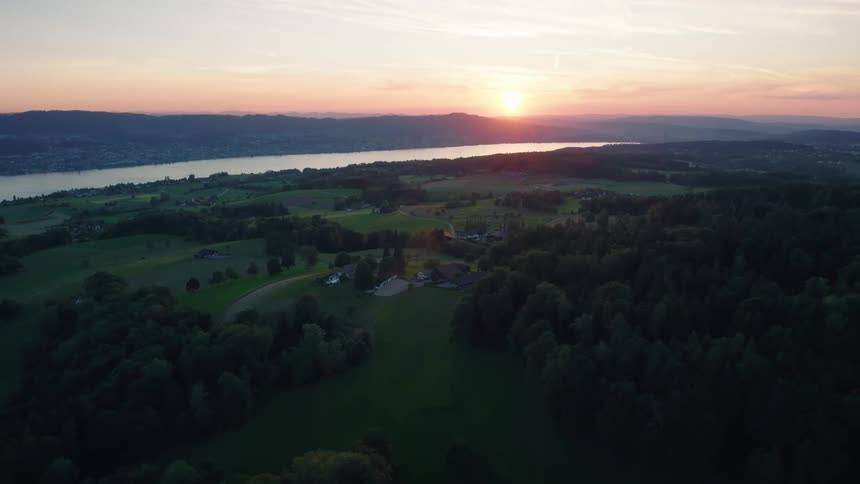 A serene aerial view shows the sun setting over a beautiful landscape of green forests and hills next to a large lake