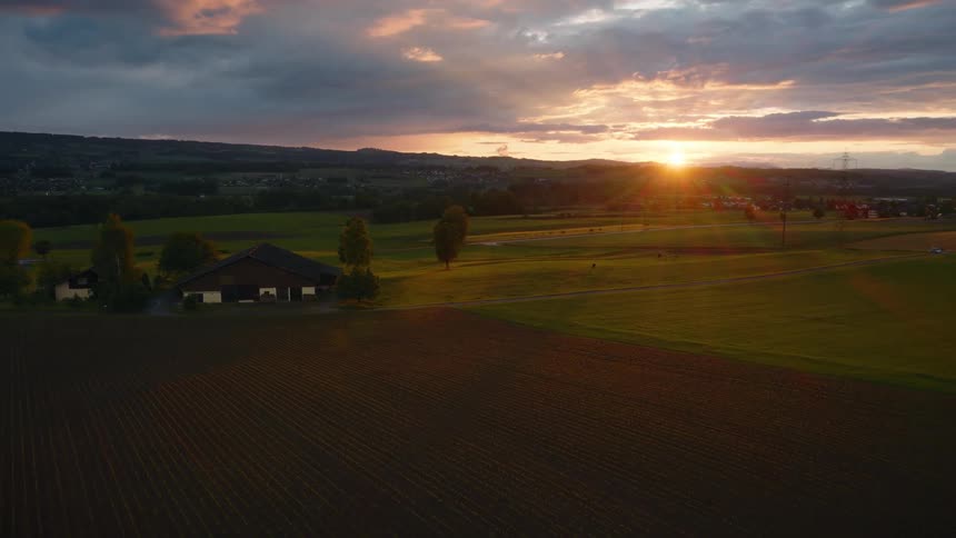 Static aerial shot of a picturesque European countryside with a farm, meadows, and plowed fields bathed in the golden light of sunset