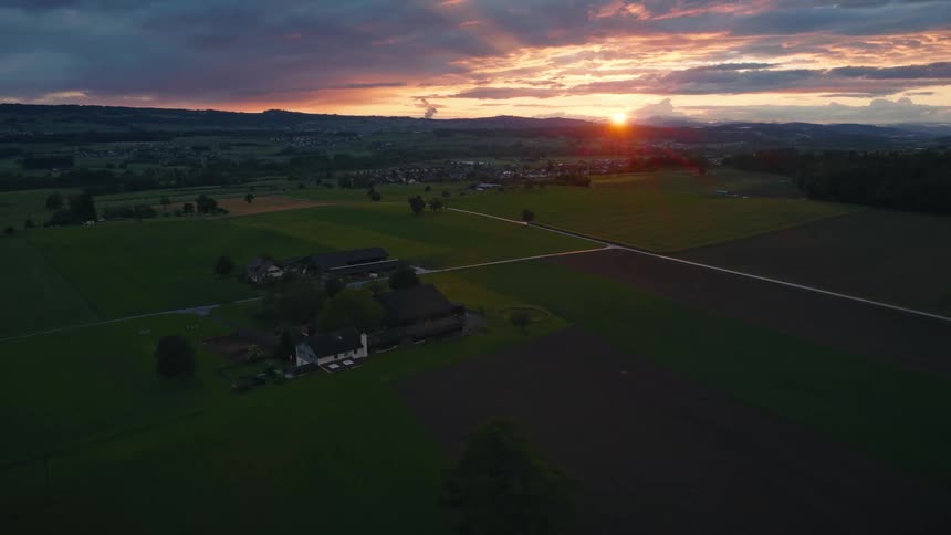 A stunning aerial view captures a farm and rolling green fields under a dramatic, colorful sunset sky