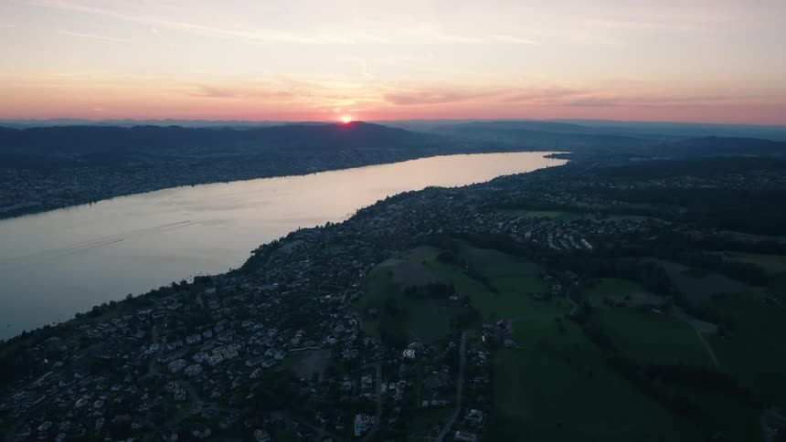 High-altitude aerial shot captures the stunning sunset over a vast lake, nestled between rolling green landscapes and lakeside communities