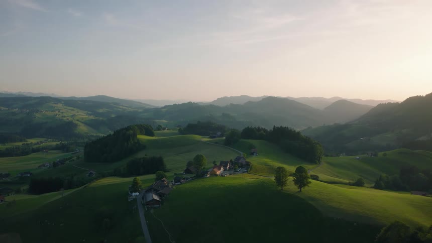A serene, static aerial shot captures the breathtaking beauty of rolling green hills and a quaint village in the countryside at sunrise