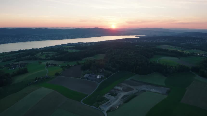 A stunning aerial view captures the serene landscape of rolling hills and patchwork farms beside a tranquil lake at sunset