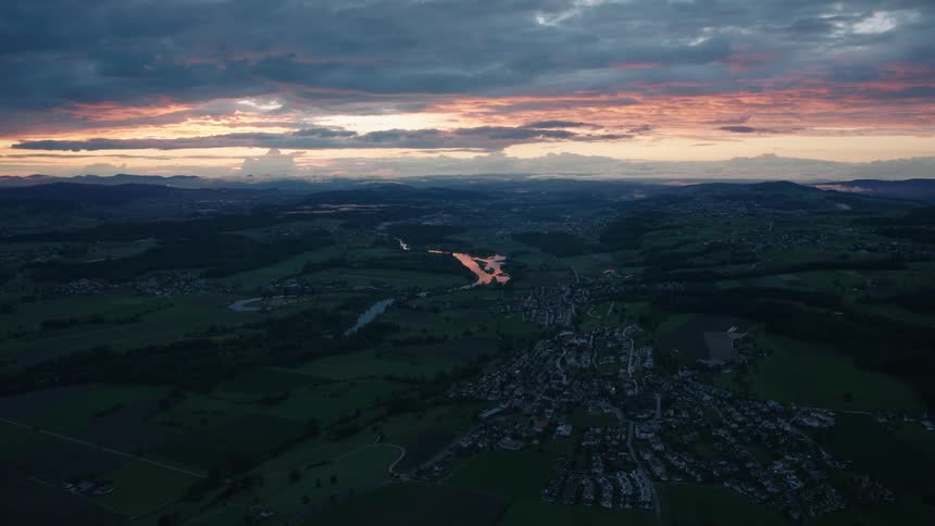 A stunning high-angle aerial view captures a tranquil town and a reflective river nestled in a vast green valley during a beautiful sunset