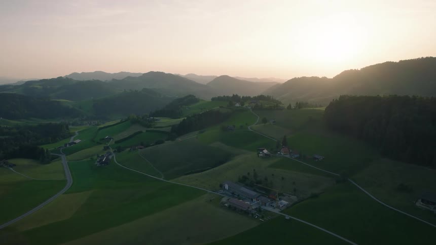 A serene aerial view flying over a vast rural landscape of rolling green hills and farms at sunrise