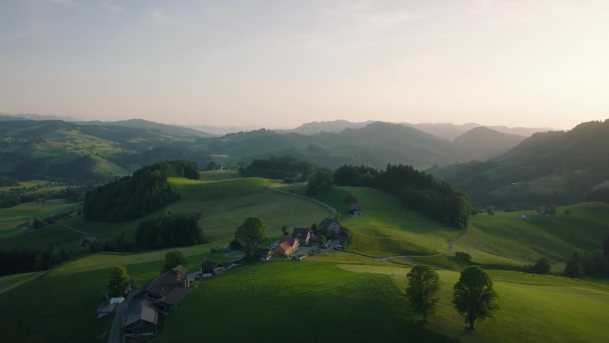 A slow aerial push-in over a small village nestled among lush, rolling green hills as the sun sets over the mountains in the distance