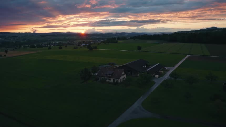 A tranquil aerial shot slowly approaches a remote farmhouse surrounded by vast green fields under a magnificent sunset sky