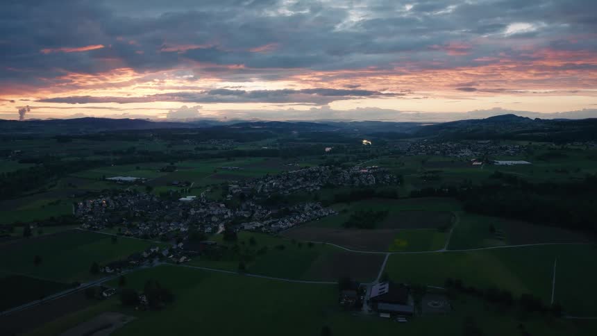 A serene aerial view shows a beautiful pink and orange sunset over a small European town and the surrounding green countryside