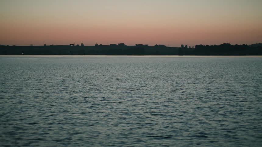 A camera tilts up from the dark, rippling water of a lake to reveal the distant shoreline silhouetted against a beautiful twilight sky