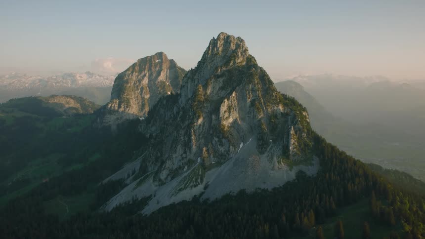 A majestic mountain peak stands tall against a hazy morning sky, with lush green forests at its base and a vast mountain range in the distance