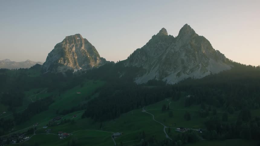 A stunning wide aerial shot shows majestic mountain peaks towering over a lush green valley and forest at sunrise