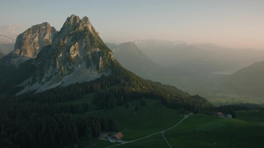 A serene static aerial view of a majestic mountain peak towering over lush green hills and a distant valley at sunrise