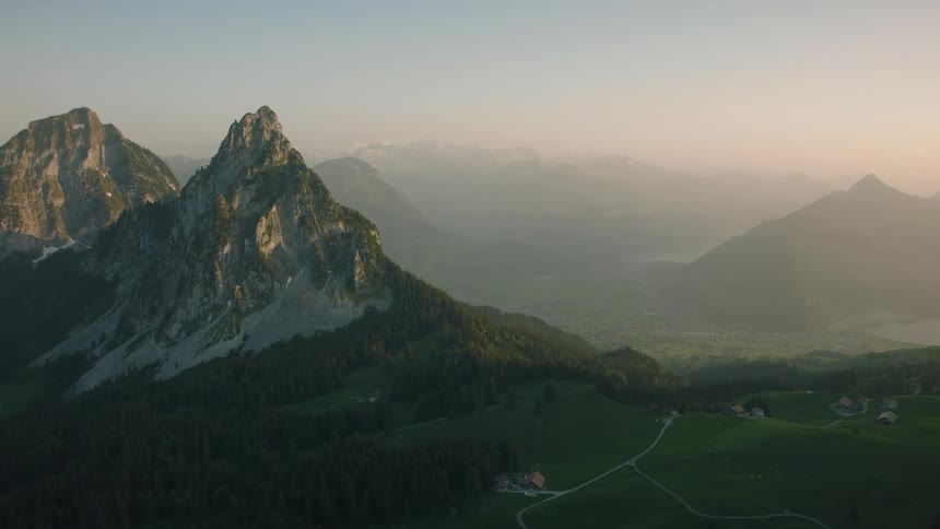 A serene, wide aerial shot of a majestic mountain range overlooking a lush green valley and forest during a hazy sunrise