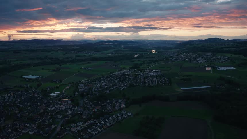 An epic drone shot flies over the Swiss countryside, showing towns and fields under a dramatic, colorful sky after sunset
