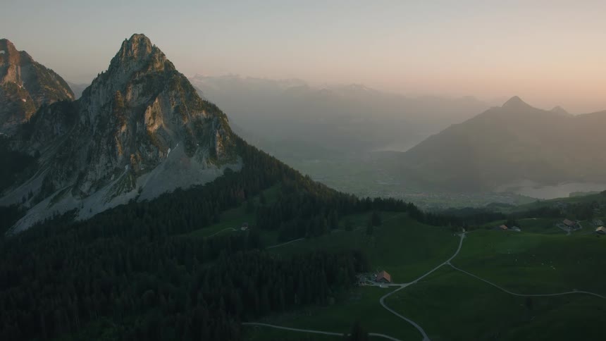A majestic mountain peak towers over rolling green hills and a hazy valley during a serene sunrise