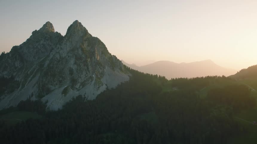 An aerial view pulls back from a majestic twin-peaked mountain and lush forest valley during a hazy golden hour sunset