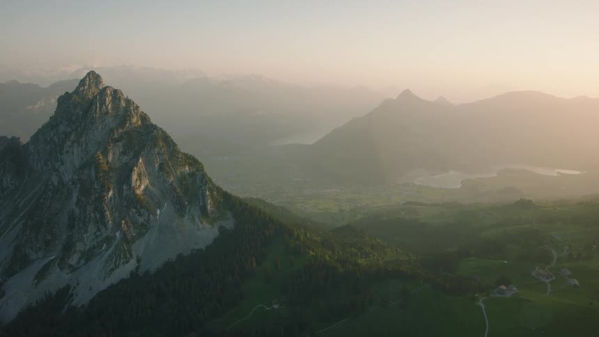A breathtaking aerial shot slowly reveals a stunning mountain range and lush green valley with distant lakes during a hazy golden hour sunrise