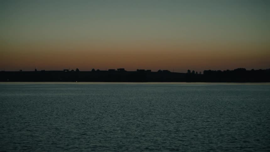 A slow tilt up from dark rippling water reveals a serene lake at dusk with a silhouetted shoreline