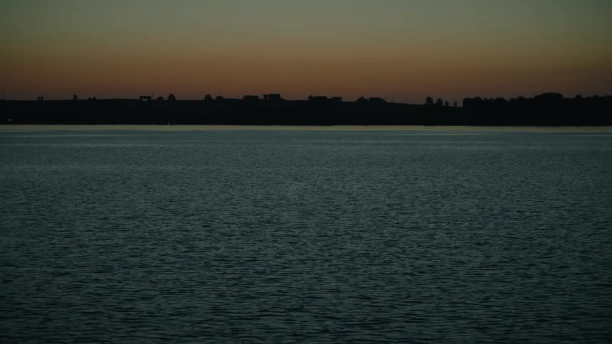 Camera tilts up over a calm, rippling lake at dusk, revealing a silhouetted shoreline against a fading orange sky