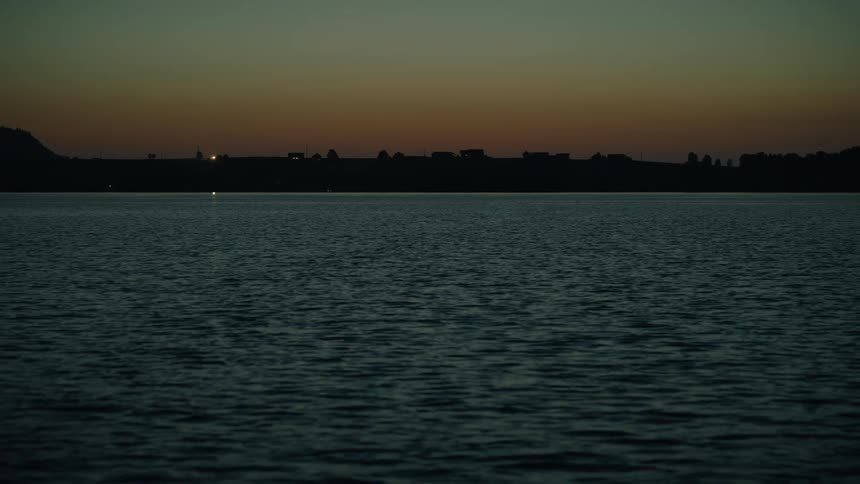 A slow tilt up from dark, rippling water reveals a calm lake and the last light of dusk on the horizon
