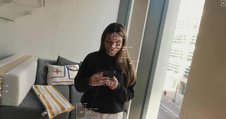 High-tech surveillance footage with data overlays tracks a woman as she scrolls on her smartphone in a modern apartment