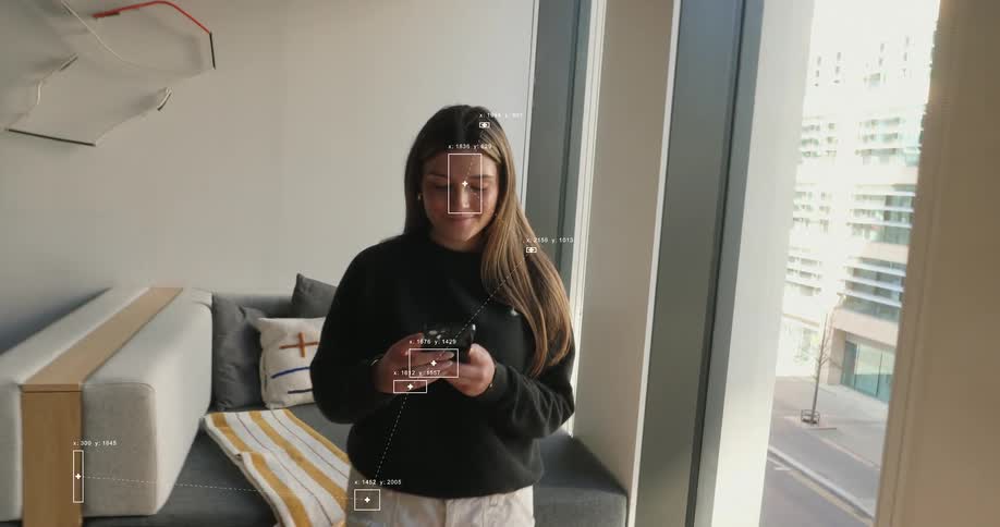 A smiling woman uses her smartphone in a modern apartment, observed by facial recognition and data analytics technology
