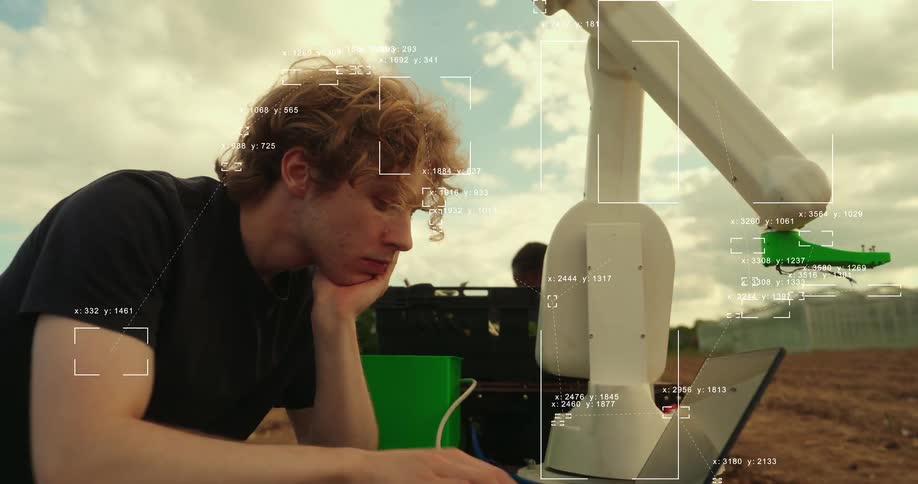 A young engineer programs an agricultural robot on his laptop as AI tracking data is displayed on screen