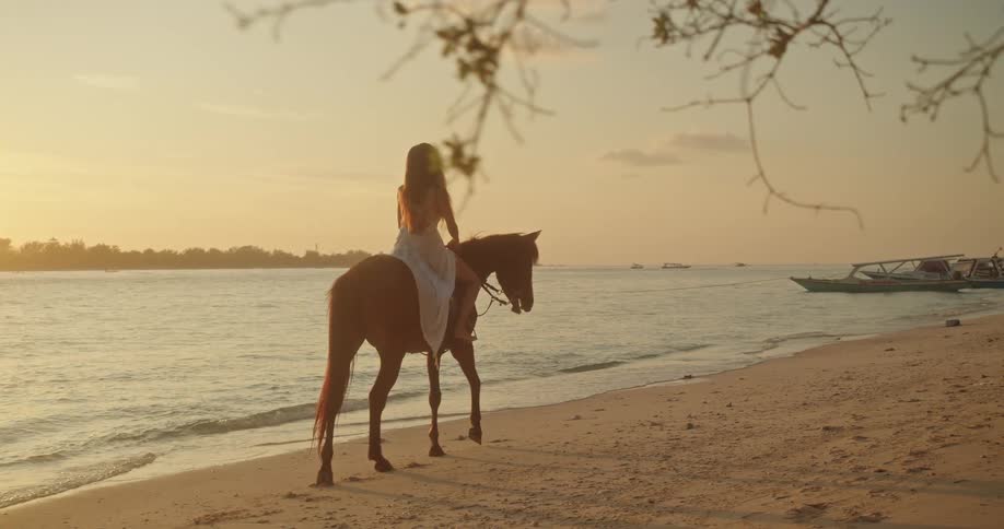 A woman in a white dress rides a brown horse along the sandy shore of a tropical beach during a golden sunset
