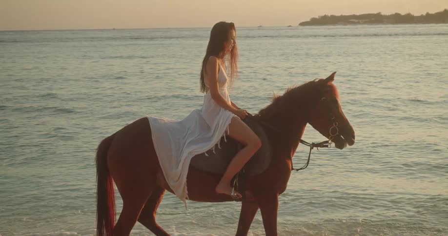 An elegant woman in a flowing white dress rides a horse along the ocean shore at sunset in slow motion