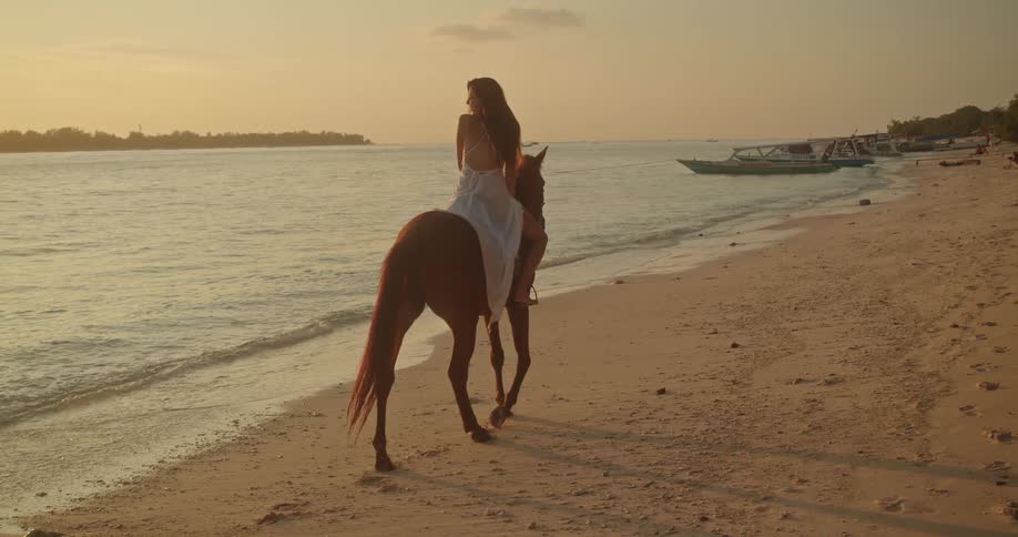 A woman in a flowing white dress enjoys a serene horseback ride along a sandy beach at golden hour in slow motion