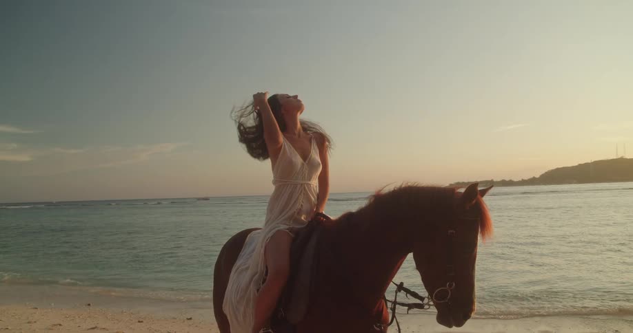 A serene woman in a white dress poses atop a beautiful brown horse on a sandy beach, bathed in the golden light of sunset