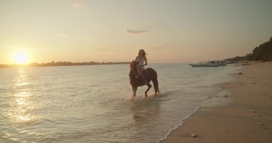 A beautiful young woman rides a horse along the sandy shore through the gentle ocean waves during a stunning golden sunset