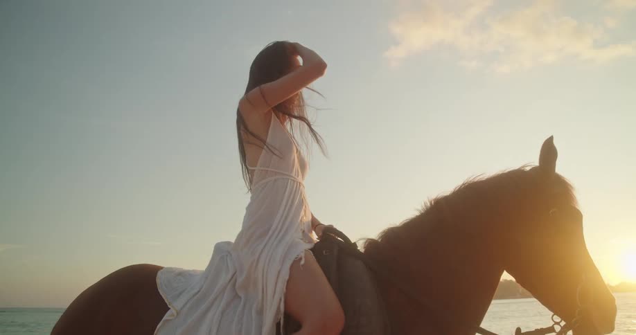 A woman in a beautiful white dress sits atop her horse on a tropical beach, enjoying the serene sunset over the ocean