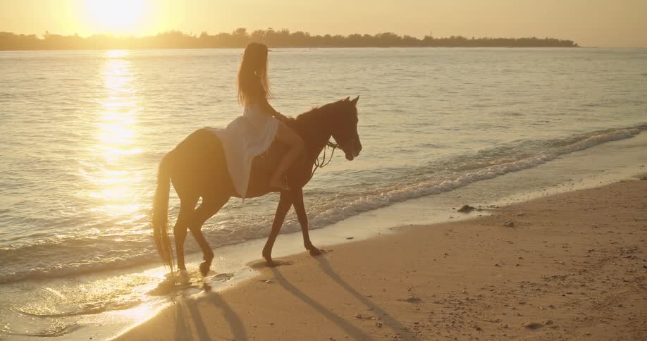 A woman wearing a white dress enjoys a peaceful horseback ride on a sandy beach at sunset