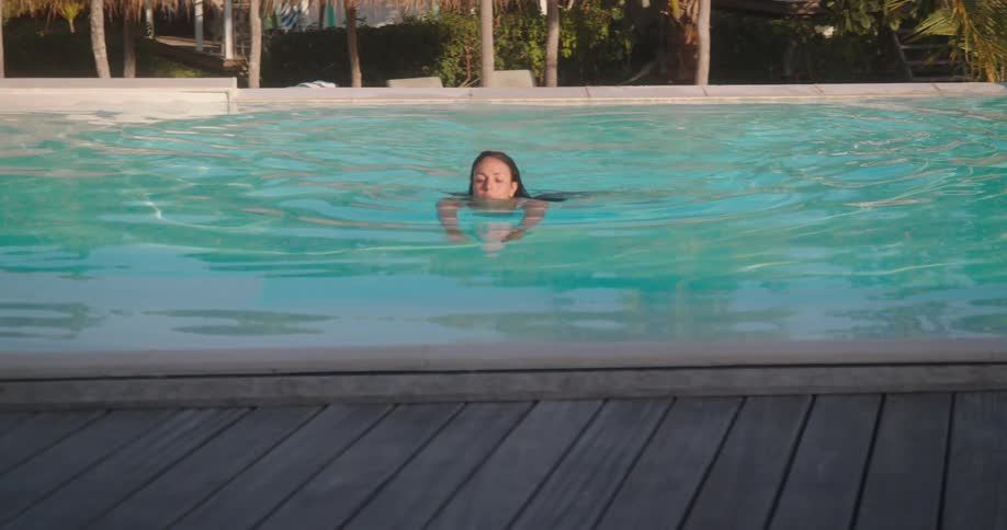 A woman peacefully swims in a tranquil turquoise pool at a tropical resort as the camera slowly pulls back