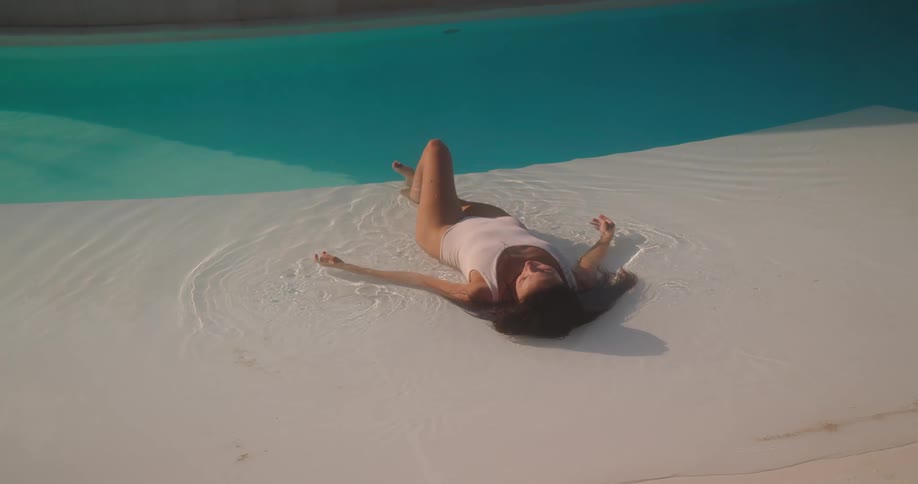 A serene woman in a white swimsuit relaxes in the shallow, sun-drenched water of a modern swimming pool as the camera pans out