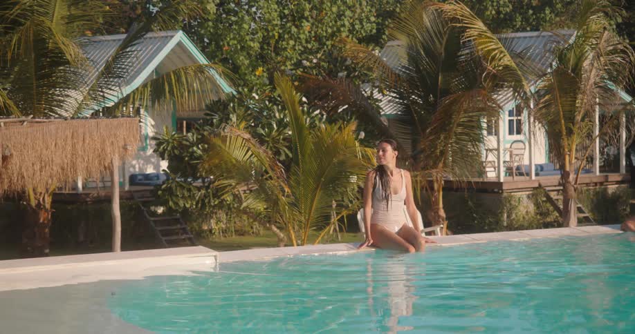A serene woman in a white swimsuit sits on the edge of a crystal clear pool, enjoying the sun at a beautiful tropical resort