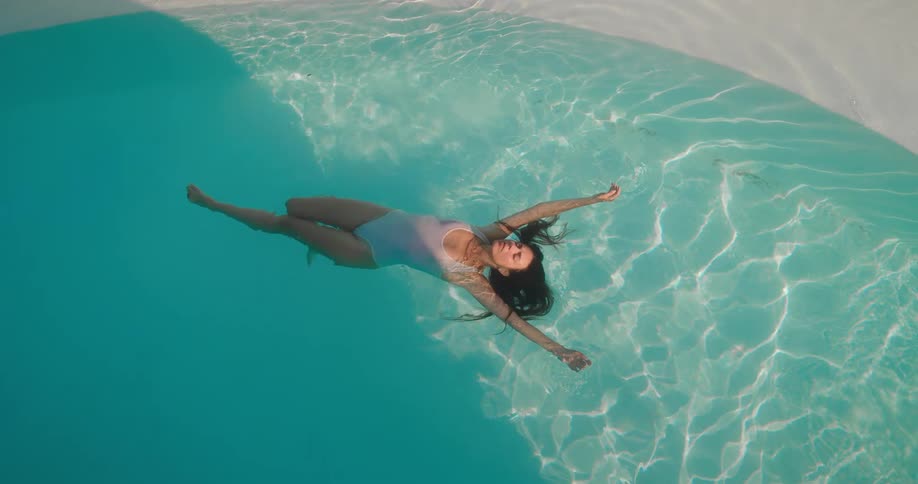 Serene top-down view of a woman in a white swimsuit floating peacefully in a crystal clear turquoise swimming pool on a sunny day