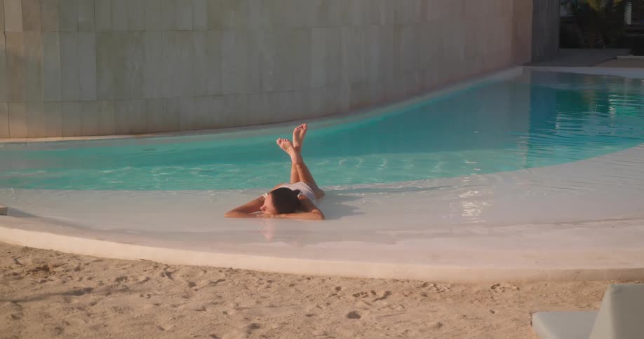 A serene woman in a white swimsuit relaxes in the shallow end of a luxurious tropical pool with a sandy beach entry