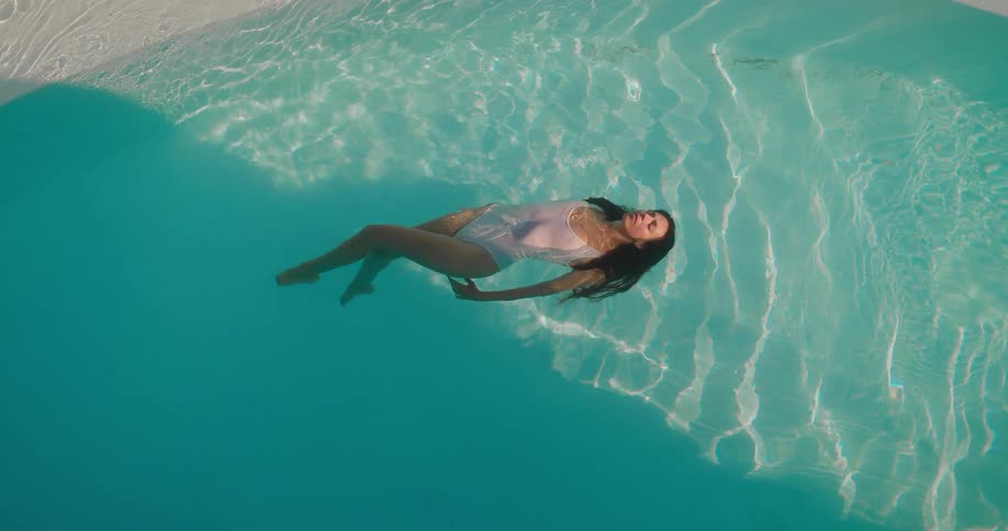 A serene overhead shot shows a woman in a white swimsuit floating peacefully on her back in a tranquil, sunlit swimming pool