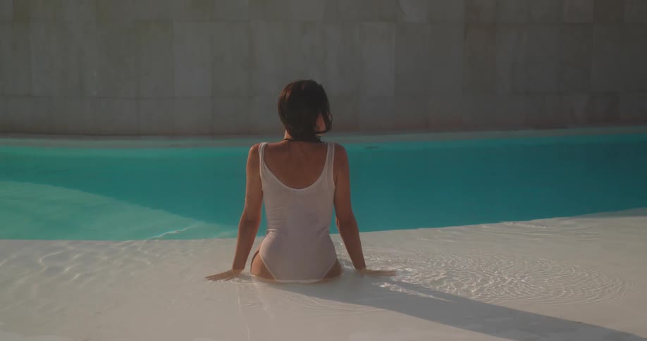 A woman with long dark hair wearing a white bathing suit relaxes in a tranquil sunlit swimming pool