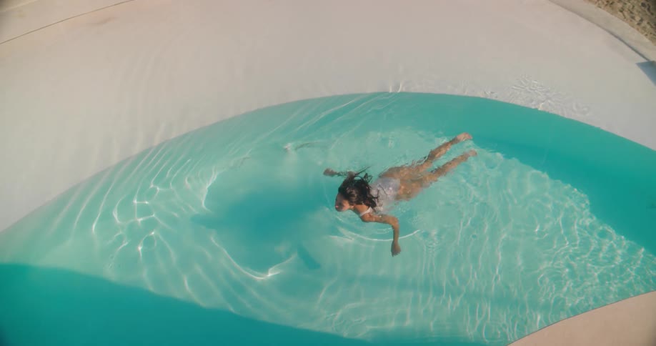 A serene woman in a white swimsuit gracefully swims in a beautiful, modern turquoise swimming pool, filmed in slow motion from above