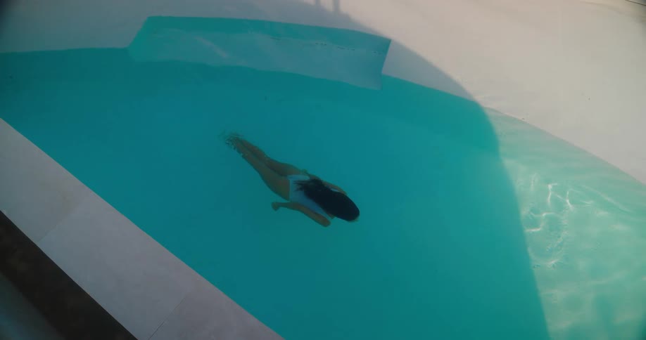 High-angle slow-motion shot pans down to a woman in a white swimsuit floating and swimming in the turquoise pool of a tropical resort