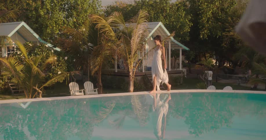 An elegant woman in a flowing white dress gracefully walks along the edge of a serene swimming pool at a tropical beach resort