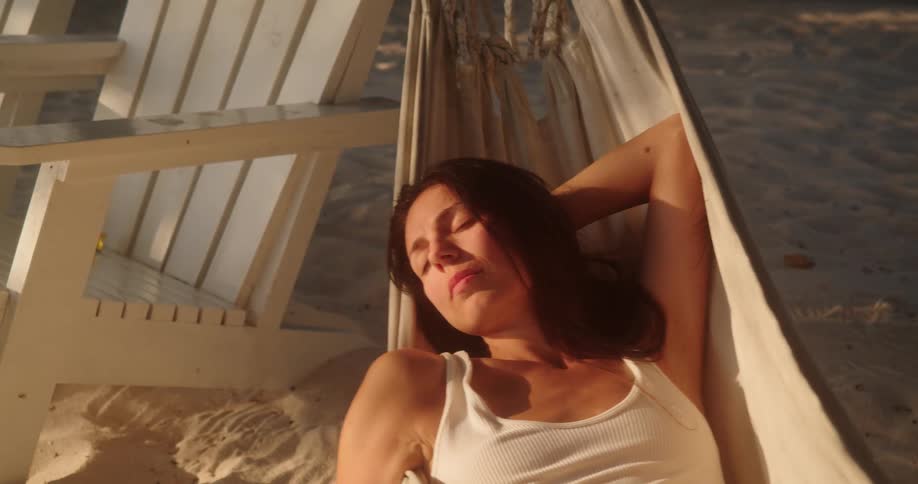 A beautiful young woman sleeps peacefully in a hammock on a sandy beach during the golden hour