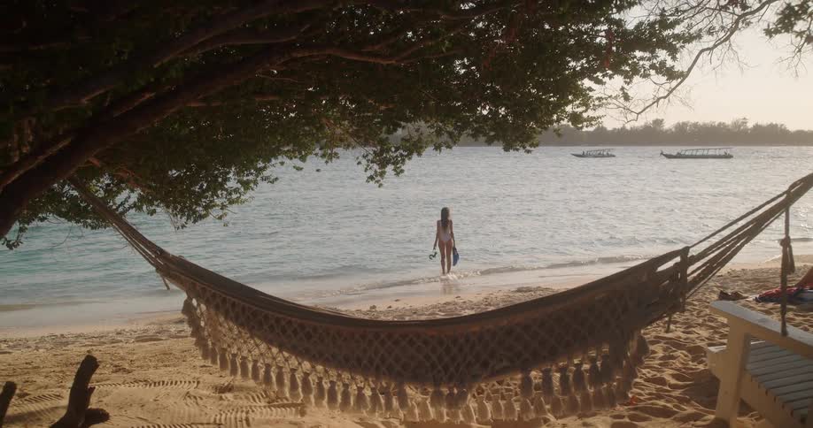 A woman with snorkel gear walks along a tranquil beach, as the camera reveals a relaxing hammock under shady trees