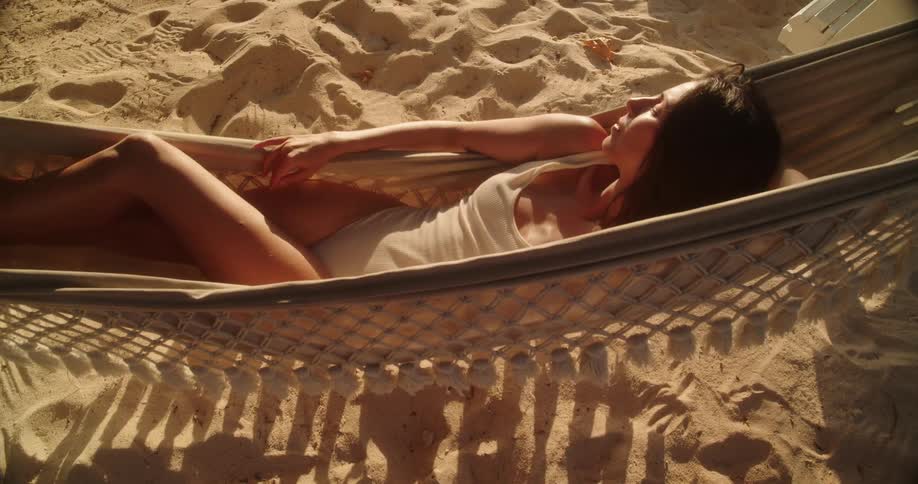 A beautiful woman in a swimsuit gently sways in a hammock on a sandy beach, enjoying the warm golden hour sunlight