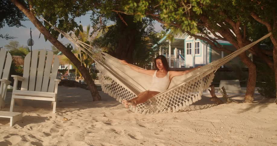 A beautiful woman in white enjoys a tranquil afternoon, swaying gently in a hammock on a sandy beach at a tropical resort