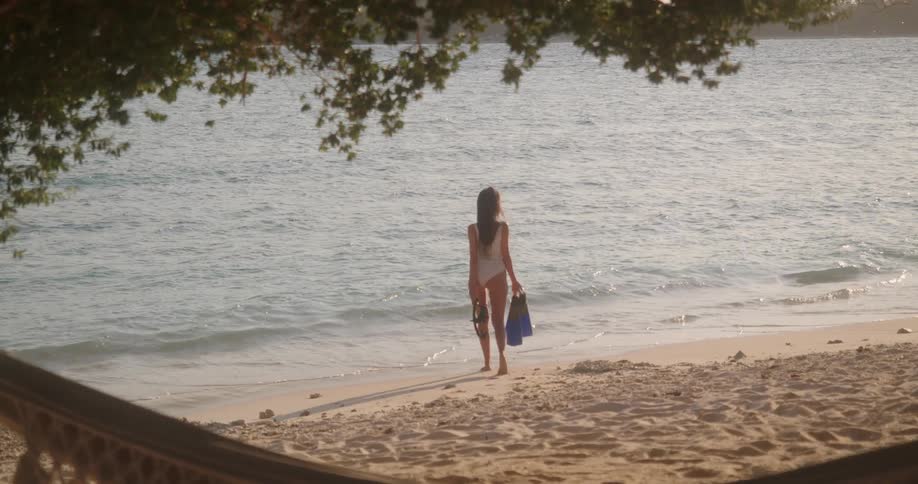 A woman in a swimsuit holding snorkel gear walks along a serene tropical beach at sunset, viewed from behind a relaxing hammock